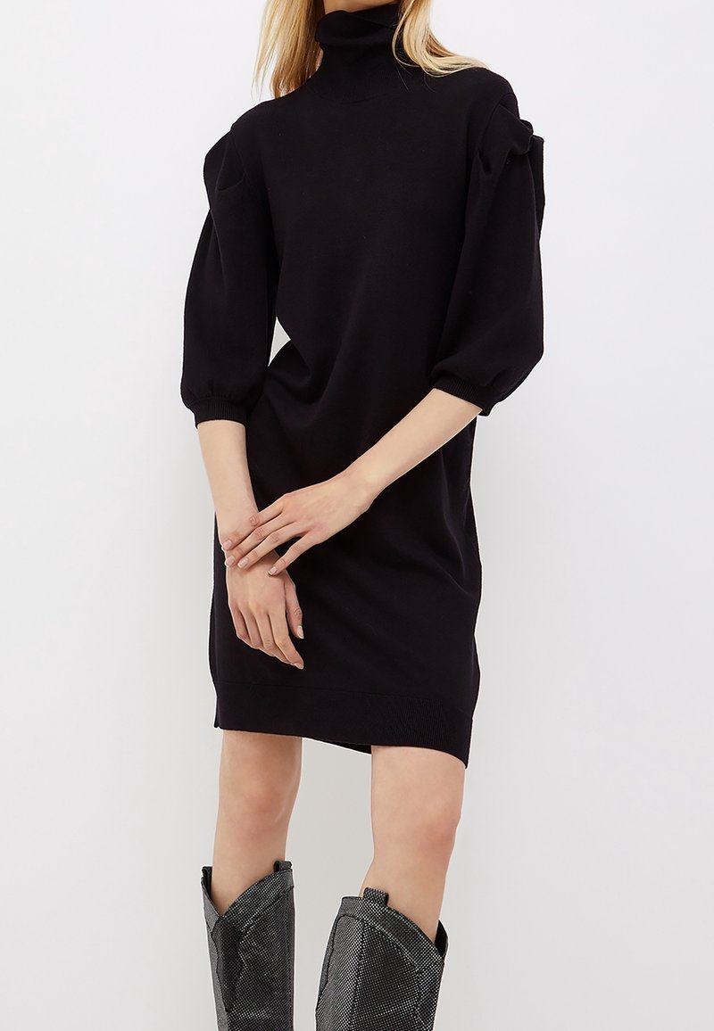 Robe en tricot noire avec un col montant, des manches ballon et une coupe décontractée, présentant des poignets côtelés et un ourlet en tissu lisse et texturé.