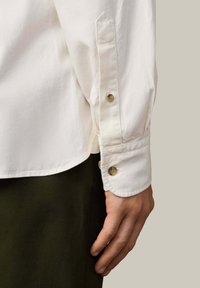 Camisa blanca de botones con dobladillo redondeado, que presenta un puño de un solo botón y una textura de algodón suave. Botones visibles en un color neutro.