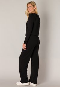 Black long-sleeve top paired with wide-leg black pants. Material appears soft with a smooth texture. Model wears white sneakers.