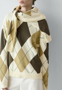 Person trägt einen beige-braun-cremefarbenen Rautenmuster-Pullover mit einem passenden Schal, der über die Schultern gelegt ist, kombiniert mit weißer Hose.