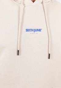 Felpa con cappuccio in beige chiaro, in tessuto testurizzato. Presenta un logo blu "SIXTH JUNE PARIS", con lacci coordinati e punte in metallo argentato.