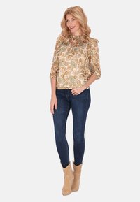Bohemian-bluse im gold-beigen Paisleymuster, mit Puffärmel und Rüschenausschnitt, kombiniert mit dunkelblauen Skinny Jeans und beigen Stiefeln.