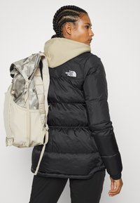 Black puffer jacket with horizontal seams, ribbed cuffs, and logo on the left shoulder; tan hoodie visible underneath; neutral backpack.