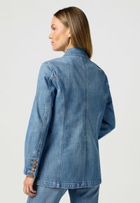 Vaaleansininen farkkutakki, jossa on muotoiltu leikkaus, lovikaulus ja napilliset hihansuut, joissa on neljä nappia. Silkkinen pinta ja takasauman yksityiskohta.