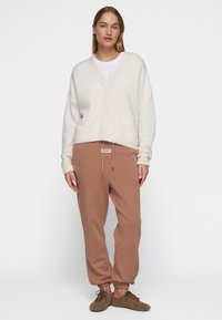 Crème gebreid trui met een V-hals, oversized pasvorm. Bruine katoenen joggers met touwtjes en geribde mouwen. Bruine veter schoenen.