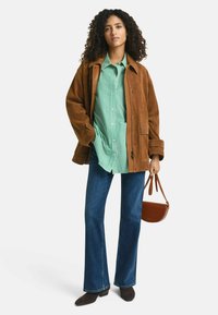 Giacca in suede marrone sopra una camicia a righe verdi e bianche, abbinata a jeans svasati blu e stivali con tacco marroni. Include una piccola borsa a tracolla marrone.