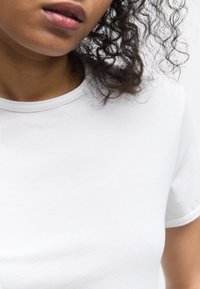 Gros plan d'une personne aux cheveux bouclés portant un t-shirt blanc uni à col rond, sur un fond clair.
