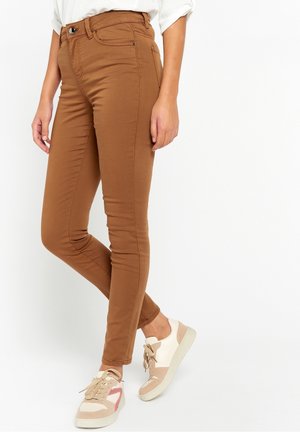 Pantalons marron ajustés en tissu lisse, dotés d'une taille haute et de poches avant, associés à des baskets de couleur claire avec des accents.