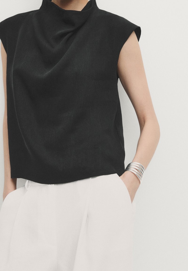 Black sleeveless top with draped neckline, made of smooth fabric. Worn with white trousers, featuring silver bangles on one wrist.