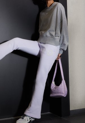 Person iført grå sweatshirt og lyse lilla joggingbukser, der holder en lavendelfarvet ternet håndtaske, løfter det ene ben mod en mørk væg som baggrund.