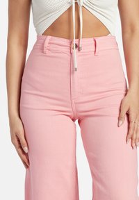 Donna che indossa pantaloni rosa a vita alta e un top corto bianco arricciato con coulisse, che mostra il ventre e ha le mani appoggiate ai fianchi.