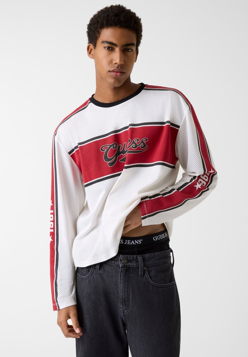 Koszulka z długim rękawem z siateczki w kolorach białym, czerwonym i czarnym z wyraźnym logo "Guess", z paskami i luźnym krojem. Noszona z ciemnymi jeansami.