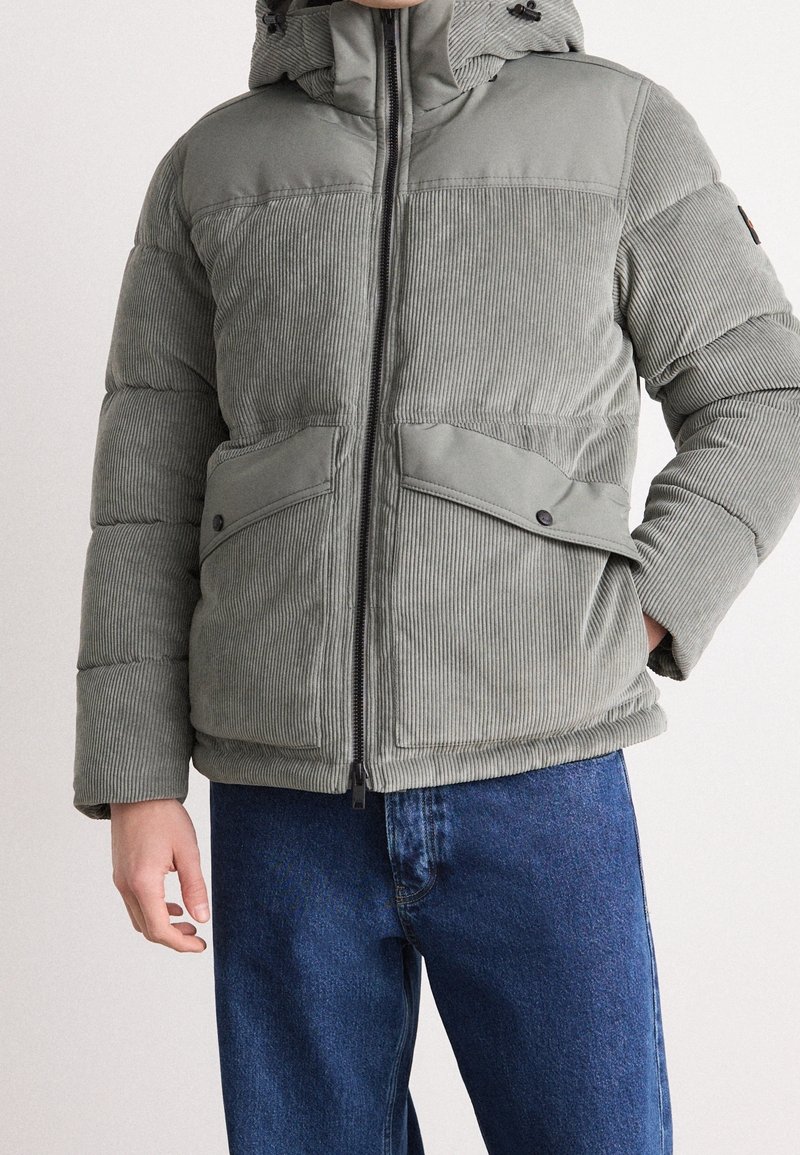 Jasnoszara pikowana kurtka sztruksowa z zamkiem z przodu, kapturem i dwoma dużymi przednimi kieszeniami. Charakteryzuje się teksturowaną tkaniną i regulowanymi paskami.