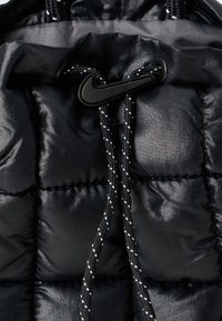 Črn puffer material z quiltano teksturo. Opremljen je s črnimi vrvicami z belimi poudarki in črnim Nike logotipom.