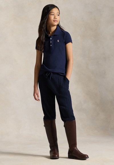 Mergina su ilgais tamsiais plaukais, vilkinti mėlynos spalvos polo marškinėlius, mėlynos spalvos pintus mezginius kelnes ir rudus aukštakulnius batus iki kelių, stovinti su rankomis kišenėse.