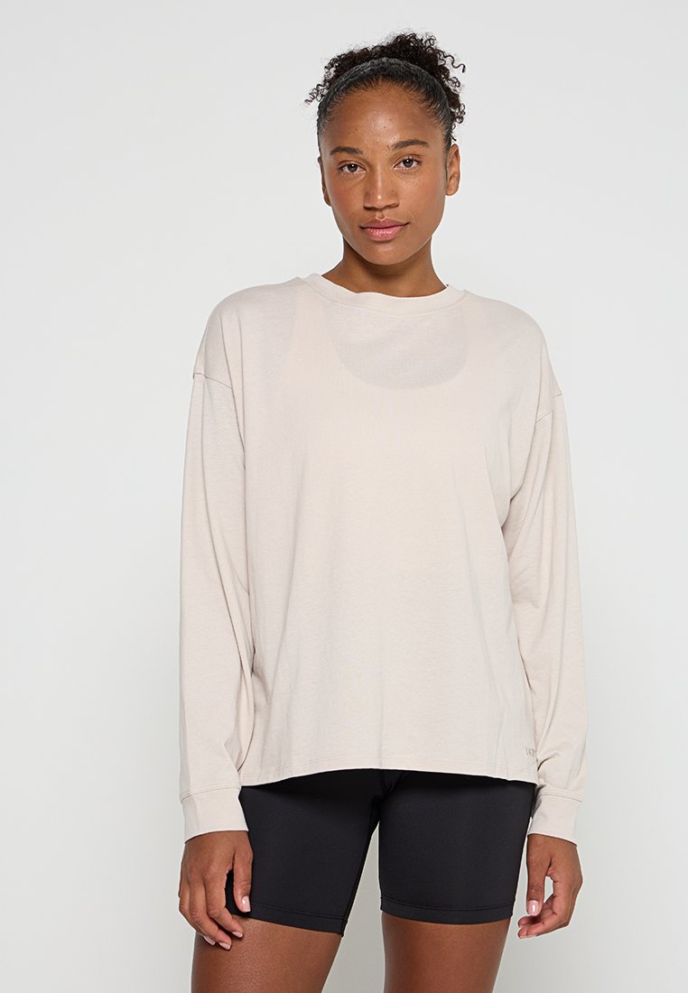 Junge Frau mit lockigem Haar, die ein leicht beiges, übergroßes Langarmshirt und schwarze, enganliegende Shorts trägt, steht vor einem schlichten Hintergrund.