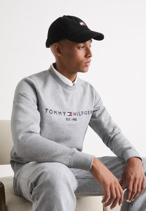 Pilkas džemperis su "TOMMY HILFIGER" logotipu, raštuotomis rankovėmis, derinamas su šviesiai pilkais džinsais. Juodas velveto skrybelė su logotipo detale. Sėdima poza.