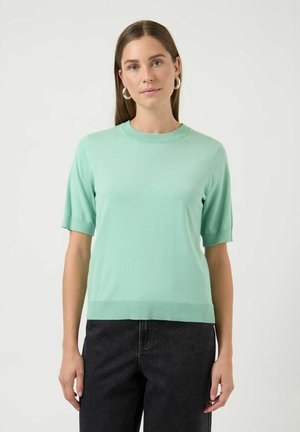 Femme aux longs cheveux bruns portant des boucles d'oreilles argentées en forme d'anneau, un pull à manches courtes vert menthe et un pantalon noir, sur un fond uni.