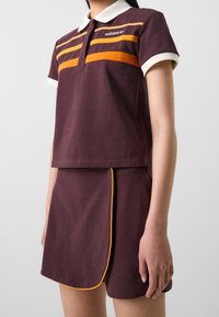 Viininpunainen poolopaita valkoisella kauluksella, jossa on oransseja vaakaraitoja ja logo. Yhdistetty hameeseen, jonka reunassa on keltaista kantta kontrastia varten.