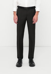 Homme portant un pantalon de costume noir slim, des chaussures en cuir noires et une chemise blanche à boutons, se tenant devant un fond uni.
