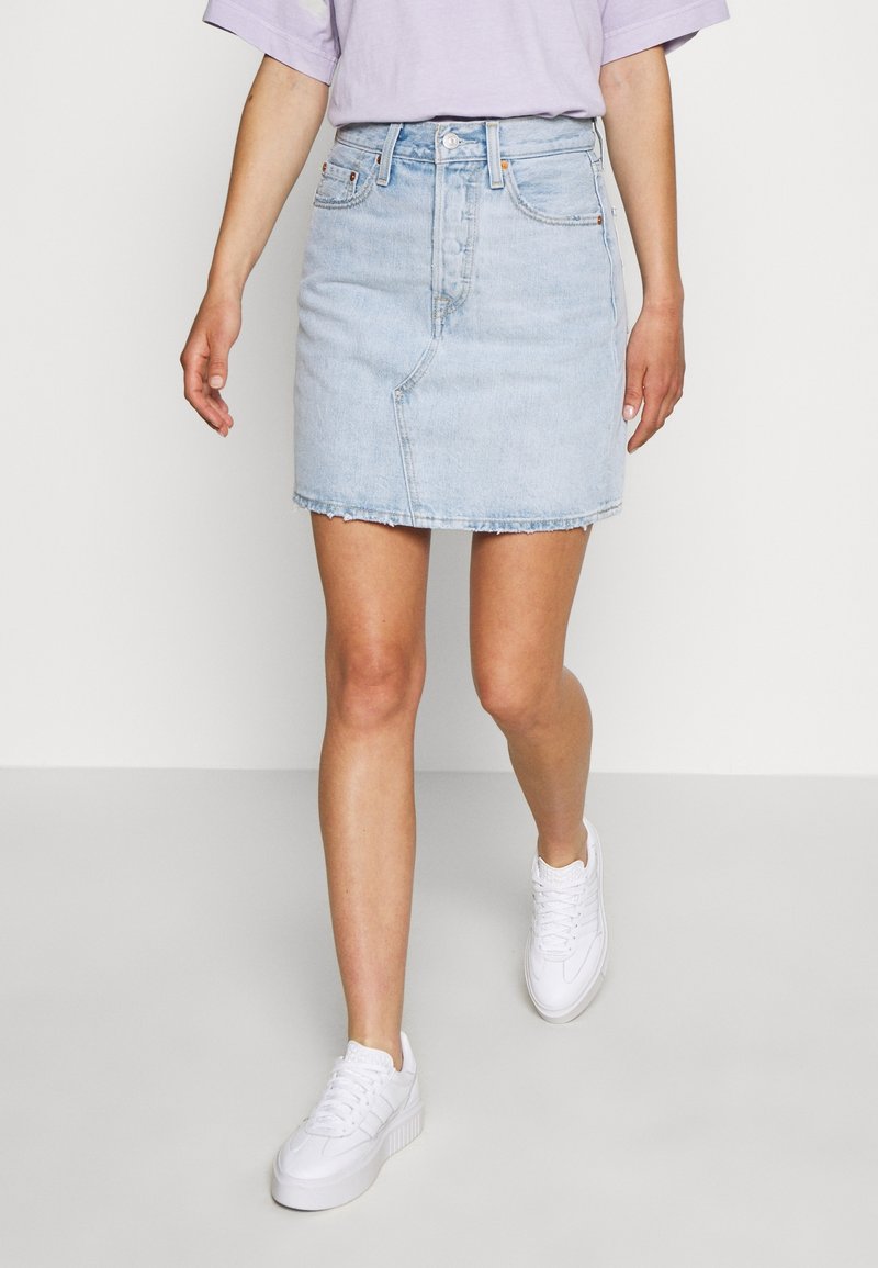 Gonna di jeans azzurri con orlo sfrangiato, forma aderente e chiusura con bottoni frontali, indossata con una t-shirt larga color lilla pastello e sneakers bianche.