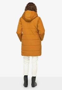 Geel pufferjack met een capuchon, doorgestikte ontwerp en een middellange snit. Draagt over een witte broek en zwarte enkellaarsjes. Achteraanzicht weergegeven.