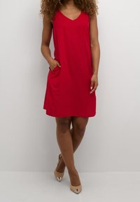 Rotes ärmelloses Kleid mit V-Ausschnitt, seitlichen Taschen und lockerer Passform, kombiniert mit beigefarbenen spitz zulaufenden Absatzschuhen. Glatte Stoffoberfläche.