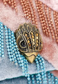 Spilla in bronzo a forma di uccello con occhio nero, adornata da strass dorati e color ambra. Accentuata da fili di perline blu e rosa.