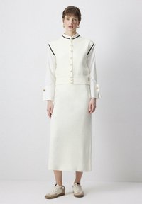 Witte gebreide vest met zwarte bies, voorzien van een rij knopen, gecombineerd met een bijpassende lange rok en beige sneakers met een gestructureerd ontwerp.