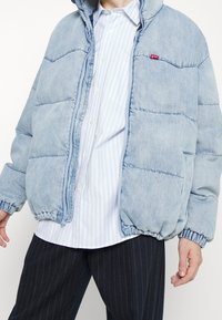 Doudoune en jean bleu clair avec capuche, poignets côtelés, fermeture éclair et poches frontales. Portée sur une chemise rayée.