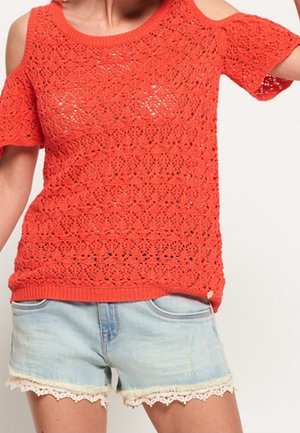 Czerwony szydełkowy top z odkrytymi ramionami, noszony z jasnoniebieskimi szortami dżinsowymi z koronkowym wykończeniem u dołu.