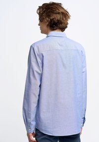 Camisa de manga larga azul claro hecha de tela chambray, con un collar tradicional y puños abotonados, y detalle de yugo en la parte posterior.