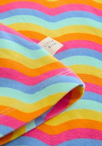 Színes szövet hullámokkal rózsaszín, narancssárga, sárga, világoskék és zöld színekben. Egy kis szövött címke az "Little Bird" felirattal az él közelében.