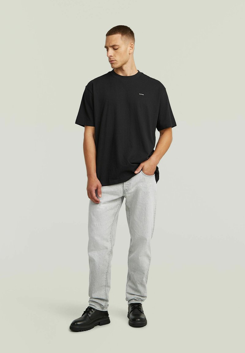 Uomo con capelli corti che indossa una maglietta nera larga, jeans grigio chiaro e scarpe nere con lacci, in piedi con una mano in tasca.