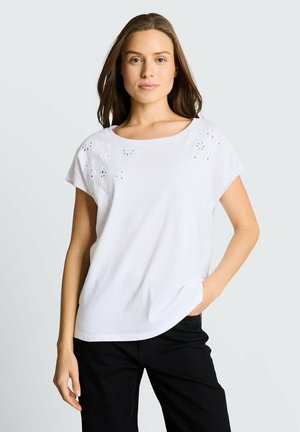 Junge Frau mit langen braunen Haaren, die eine weiße Kurzarmbluse mit floralen Ausschnitten und schwarze Hose trägt, Hand in der Tasche.