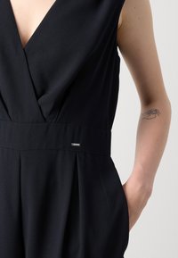 Svart ermeløs jumpsuit med dyp V-hals, plisserte detaljer i midjen, en metalllogo og en glatt tekstur.