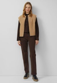 Beige, pluizig vest over een donkerbruin overhemd met knopen en bijpassende broek, gecombineerd met zwarte platform enkellaarsjes. Neutrale achtergrond.