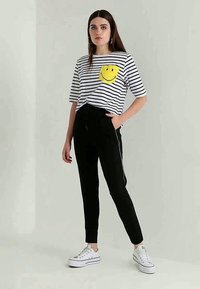 T-shirt às riscas brancas e pretas com um sorriso amarelo no bolso, combinada com calças pretas e sapatilhas brancas; look casual.