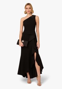 Czarna sukienka na jedno ramię z falbaną u dołu, z wysokim rozcięciem. Gładka tekstura z koralikowym akcentem na ramieniu. Modelka trzyma czarną kopertówkę.