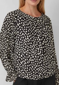 Schwarze Bluse mit unregelmäßigen weißen Punkten, Rundhalsausschnitt und gerafftem Detail; verfügt über lange Ärmel mit Rüschensäumen. Glatte Stofftextur.