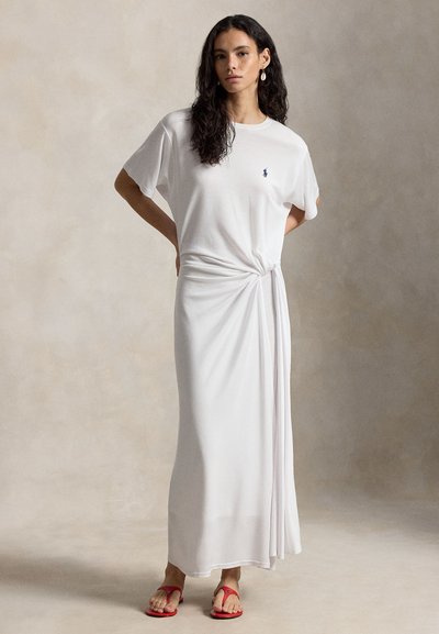 Νεαρή γυναίκα με μακριά σγουρά μαλλιά, φορώντας λευκό μακρύ φόρεμα με λεπτομέρειες δεσίματος και κόκκινες σανδάλια, στέκεται μπροστά σε απλό μπεζ φόντο.