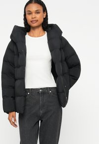 Čierna prešívaná bunda s kapucňou, so zipsom vpredu a bočnými vreckami, nosená cez bielu košeľu a tmavé džínsy.