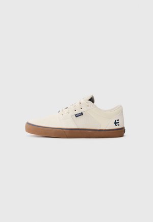 Low-top beige canvas sneaker with brown rubber sole, lace-up front, and small branded tags on side and heel.
