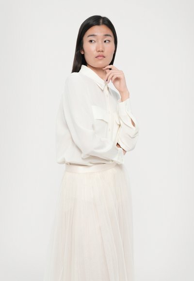Witte, lange mouwen blouse met knoopsluiting en twee voorzakken, gecombineerd met een vloeiende, doorschijnende crèmekleurige rok met een geplooide textuur.