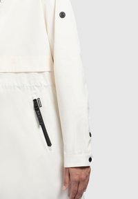 Giacca impermeabile bianca con una tasca con zip nera, dettagli a pressione e una texture liscia. La manica presenta un'etichetta con logo sulla spalla.