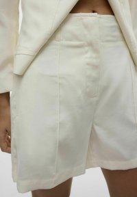 Pantaloni corti giallo chiaro con una texture liscia, dotati di tasche laterali e una vestibilità sagomata. Abbinati a una giacca coordinata per un look armonioso.