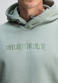 Lichtgroene hoodie met een trekkoordkap, voorzien van "BUTCHER OF BLUE" geborduurd in een iets donkerder groen. Zachte, gestructureerde stof.
