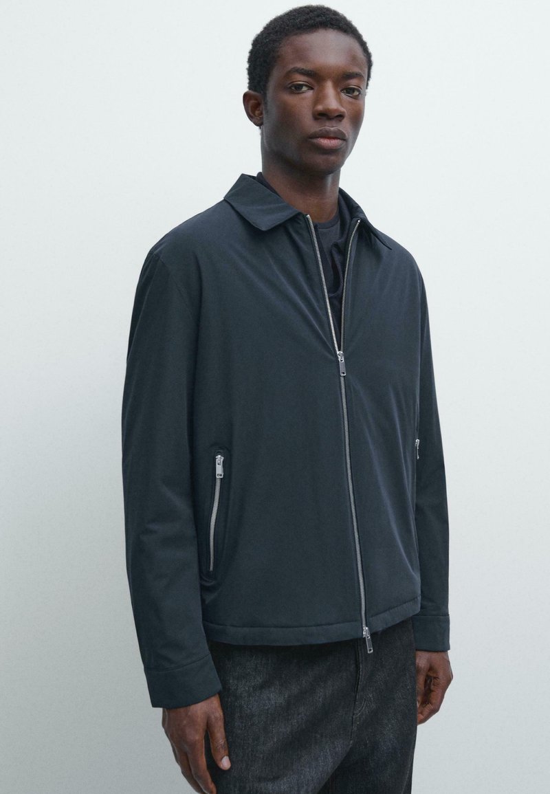 Mand iført en sort jakke med lynlås og lynlåslommer over mørke bukser, stående mod en ensartet lys baggrund.