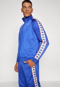 Blauwe trainingspak gemaakt van gladde stof, met een hoge kraag en rits. Zijstrepen bevatten een herhaald logo in het wit met rode accenten.