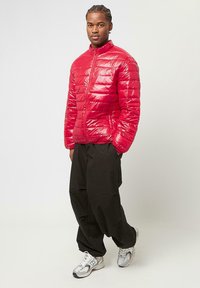 Rote Pufferjacke mit Reißverschluss, die ein gestepptes Design aufweist. Kombiniert mit lockeren schwarzen Hosen und silbernen Sportschuhen.